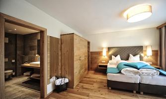 A modern hotel room with a large double bed, warm hardwood floors, and soft colors. In the background, there is an adjacent bathroom.