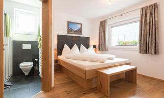 A bright bedroom with a large bed and wooden details. In the background, a bathroom in neutral colors is visible.