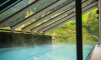 A modern pool area with a glass ceiling. In the background, green hills and mountains can be seen.