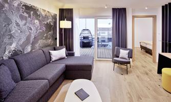 A modern living room with a gray sofa and a minimalist design. Large windows provide access to a balcony and plenty of natural light.