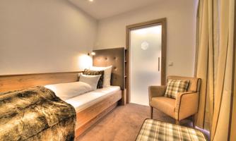 A simple bedroom with a single bed and a cozy armchair. The room has bright light and a relaxed atmosphere.