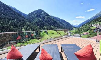 A cozy balcony with a view of green hills and majestic mountains. Red cushions on modern lounge furniture invite relaxation.