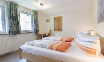 A cozy bedroom with two beds and colorful pillows. Large windows provide plenty of daylight and an inviting space.