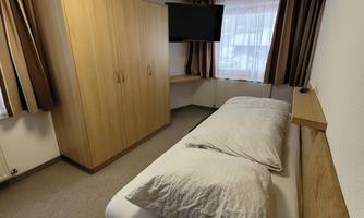 A simple room with a single bed and a television. The walls are light and there are windows with curtains.