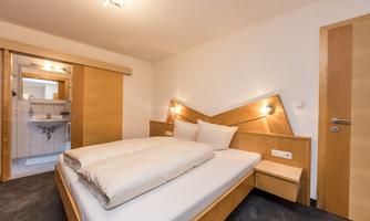 A modern bedroom with a large double bed and light wooden furniture. In the background, a bathroom with access is visible.