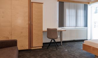A simple room with a desk, a chair, and a wardrobe. The window has blinds and lets in a lot of light.