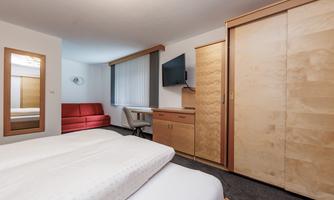 A modern room with a double bed and a television. In the background, there is a red sofa and a wooden wardrobe.