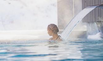A person is relaxing in the water of an outdoor pool. In the background, snow-covered landscapes and buildings can be seen.