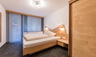 A modern bedroom with a double bed and light wooden furniture. Large windows with blinds provide plenty of daylight.