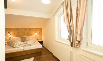 A cozy bedroom with a large bed and warm wood details. The room has bright windows and soft curtains.