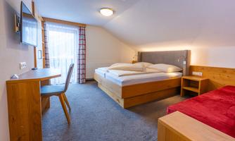 A modern hotel room with a double bed and a single bed. There is a desk, a television, and large windows with a view outside.