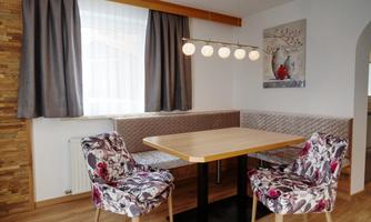 A cozy dining area with a table and two chairs. The walls are light, and there are large windows with curtains as well as decorative wall art.