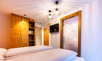 A modern bedroom with a large bed and a wooden chest of drawers. The walls are light, and there is stylish lighting.