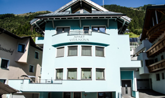 A modern building in light blue color with large windows. The house is named "Apart Vita Nova" and is located in a picturesque setting.