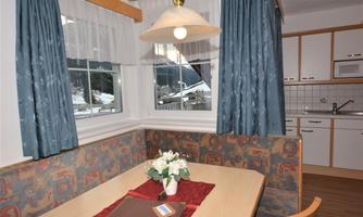 A bright dining area with a table and a cozy bench. Blue curtains hang in front of a window, and a flower arrangement sits on the table.