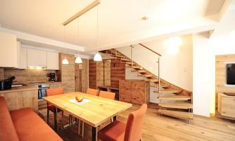 A modern living room with a wooden staircase and dining area. The decor is bright and inviting with warm wooden tones and an open kitchen.