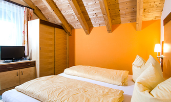 A cozy room with wooden ceilings and two single beds. The walls are painted orange and there is a television as well as a wardrobe.