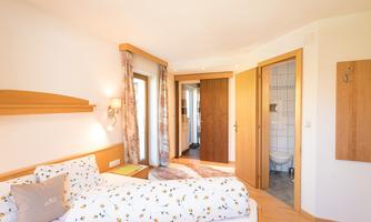 A cozy bedroom with a double bed and bright walls. The door to the bathroom is open and there are windows for daylight.