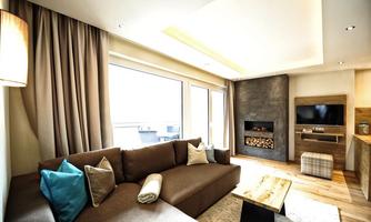 A modern living room with a large sofa, cozy cushions, and a fireplace. Large windows provide plenty of light and an inviting atmosphere.