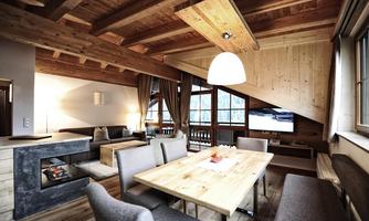 A modern, cozy living room with a wooden ceiling and large windows. The wooden dining table is at the center, surrounded by stylish chairs.