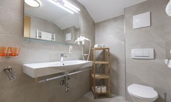 A modern bathroom with a sink, a toilet, and a wooden shelf. The walls are designed in neutral gray tones and there is soft lighting.