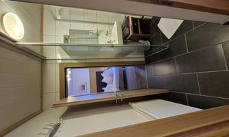 A modern bathroom with a glass shower and a sink. In the background, there is a bedroom with a double bed.
