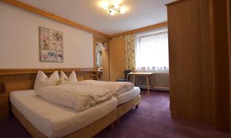 A cozy room with a double bed and wooden paneling. Bright windows let in plenty of light, and there is a small table with a chair.