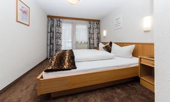 A cozy bedroom with a large bed and wood-colored furniture. The windows are decorated with curtains and there is soft lighting.