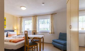 A bright room with a double bed, a table, and chairs. There are large windows that let in lots of daylight.