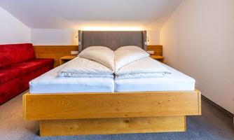 A modern bedroom with a large bed and soft pillows. In the background, there is a red sofa on a carpeted floor.
