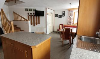 A bright kitchen with a wooden island and a dining area. In the background, a staircase and windows for natural light can be seen.