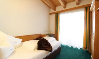A bright room with a single bed and wooden ceiling. Large windows with curtains let in plenty of daylight.