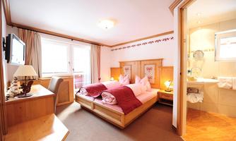 A cozy hotel room with two beds and wooden decor. In the background, there is a bathroom area and a window with plenty of daylight.