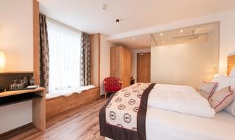 A modern bedroom with a large bed and wooden decor. Bright windows provide plenty of daylight and a cozy atmosphere.