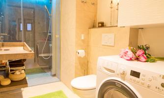 A modern bathroom with a shower and washing machine. On the shelf, there are roses and there is a green bath towel on the floor.