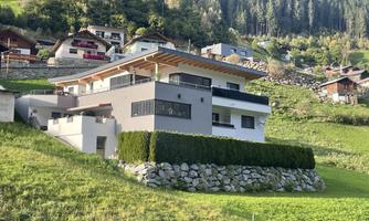 A modern house on a hill, surrounded by green areas. In the background, other houses and trees can be seen.