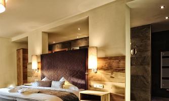 A modern bedroom with a large bed and elegant lighting. The walls are made of light wood, and a bathroom is visible.