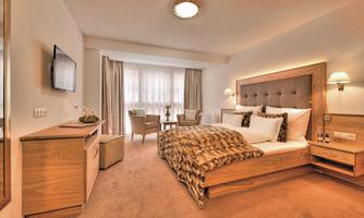 A modern bedroom with a large bed and wooden furniture. The room is bright and cozy, with a small seating area and windows that let in plenty of light.