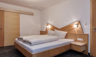 A modern bedroom with a wooden bed and two pillows. The walls are simple and the lighting is pleasant.