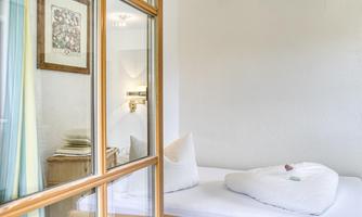 A cozy room with a single bed and white pillows. The room has windows with colorful curtains and a decorative item on the wall.