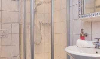A modern bathroom with a clear shower cabin and a sink. The walls are tiled and there is a mirror above the sink.