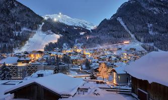 A snow-covered landscape at night with a small village and illuminated houses. In the background, mountains and ski slopes are visible.