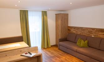 A bright living room with a cozy sofa and a wooden cabinet.  
Large windows let in plenty of light and there are green curtains.
