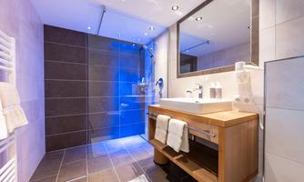 A modern bathroom with an elegant wooden bench and a large mirror. The shower is glass and there is soft lighting.