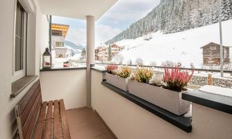 A balcony with a wooden bench and two large planters. In the background, snow-covered mountains and a quiet village can be seen.