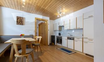 A modern kitchen with wooden ceilings and light furniture. Visible are a table, chairs, and various kitchen appliances.