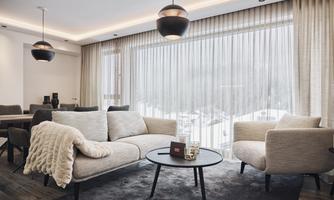 A modern living room with a comfortable, bright sofa and a round coffee table. Large windows with curtains offer a view of a wintry landscape.