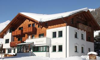 A charming wooden building in the alpine style, surrounded by snow-capped mountains. The facade combines wood and plaster surfaces.
