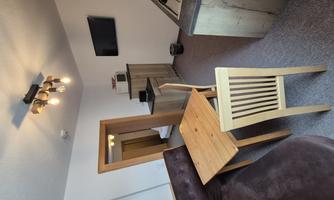 A modern living room with a couch, a wooden table, and a chair. In the background, there is a kitchen with a TV on the wall.