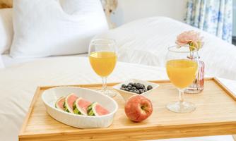 A breakfast tray on a bed, showing two glasses of juice, an apple, and a bowl of blueberries. Next to it are fresh melon slices in a white bowl.